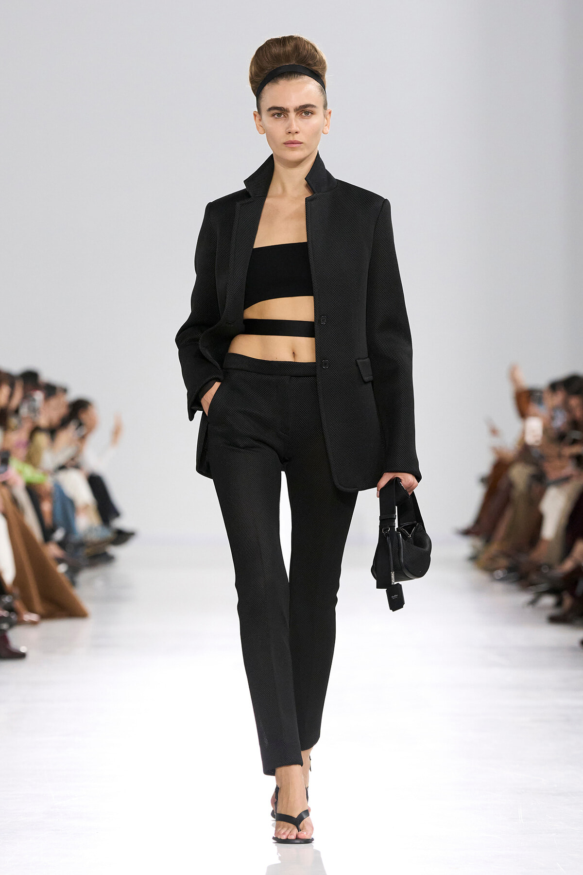 Model walking a runway in a black textured suit with a cropped black bandeau top and cutout straps, carrying a small black handbag.