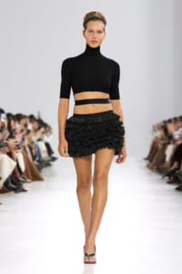 Female model walks the runway in a black turtleneck crop top and a textured black ruffled mini skirt, with a headband and sandals, audience visible on both sides.
