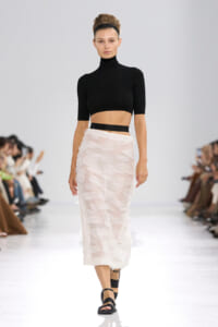 Model walking the runway in a black cropped turtleneck and pale pink textured midi skirt with a black headband and black sandals.