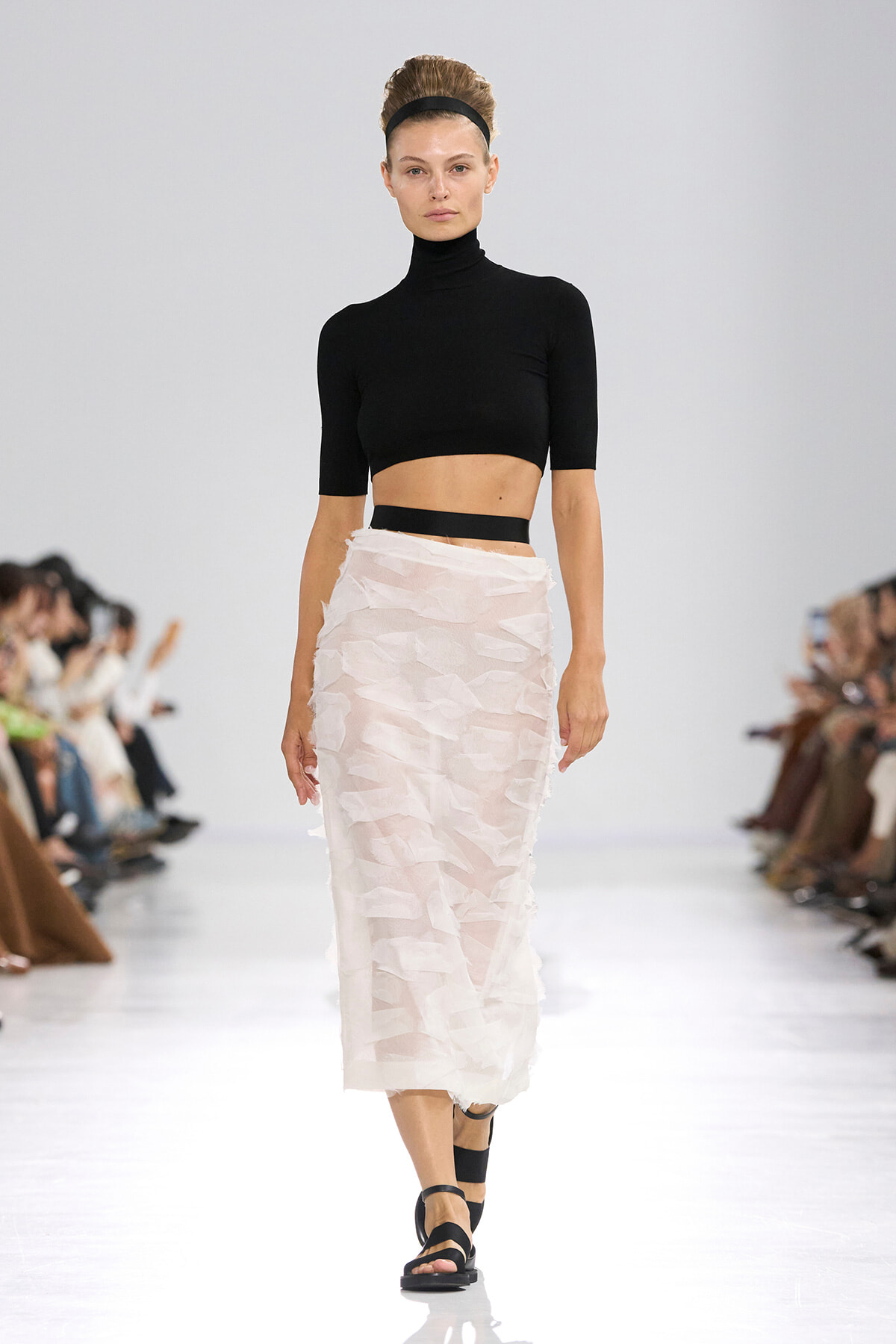 Model walking the runway in a black cropped turtleneck and pale pink textured midi skirt with a black headband and black sandals.