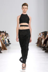 Model walking the runway in a black halter crop top and long textured skirt, with a black headband.