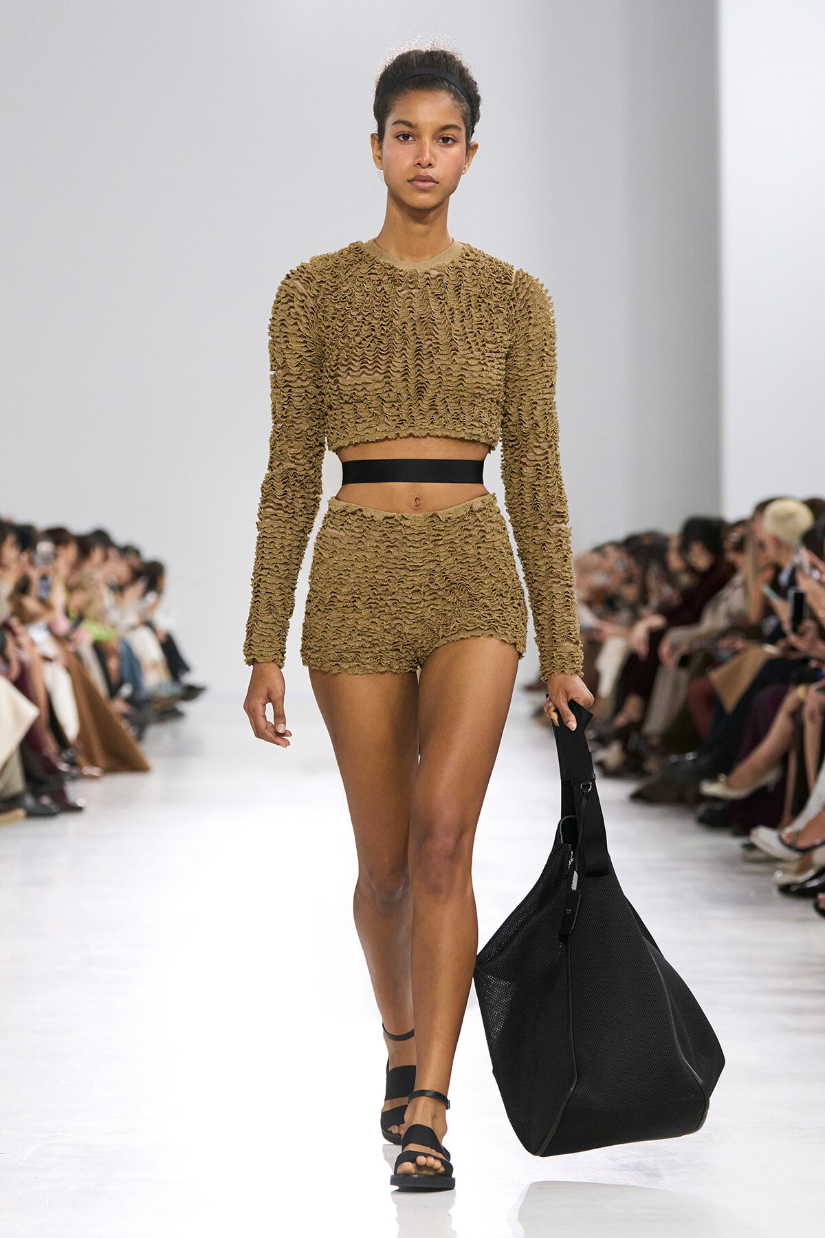 Female model walks a fashion runway in a textured tan crop top and matching shorts, carrying a large black tote bag as the audience watches.