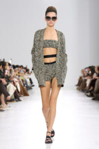 Model on a runway wearing a textured black-and-beige knit set: bandeau top, matching shorts, and an oversized cardigan with black sandals and sunglasses.