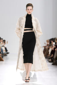 Model walking runway in a black two-piece outfit with a long beige textured coat, hair up with a headband.