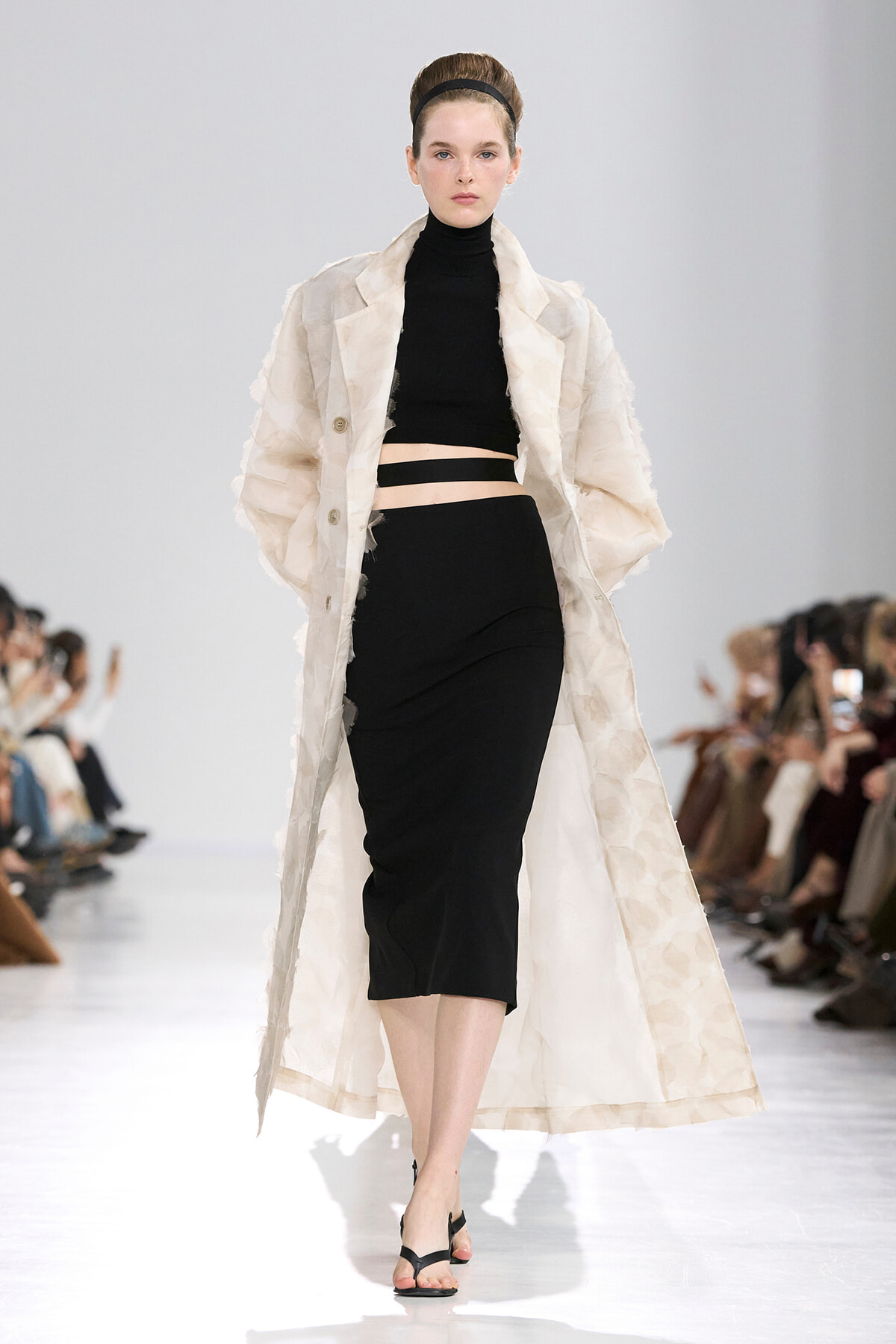 Model walking runway in a black two-piece outfit with a long beige textured coat, hair up with a headband.