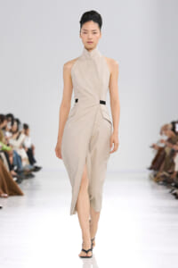 Female model walks a runway in a beige sleeveless high-neck wrap dress with a front slit and a thin black waist belt for contrast.