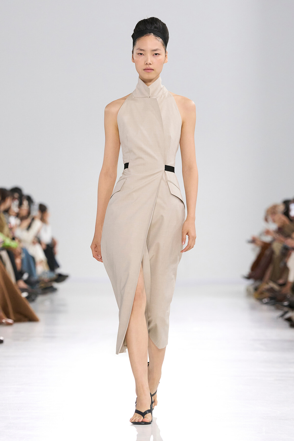 Female model walks a runway in a beige sleeveless high-neck wrap dress with a front slit and a thin black waist belt for contrast.