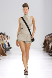 Model walks the runway in a beige sleeveless tailored romper with a high collar, paired with a black crossbody bag and headband.
