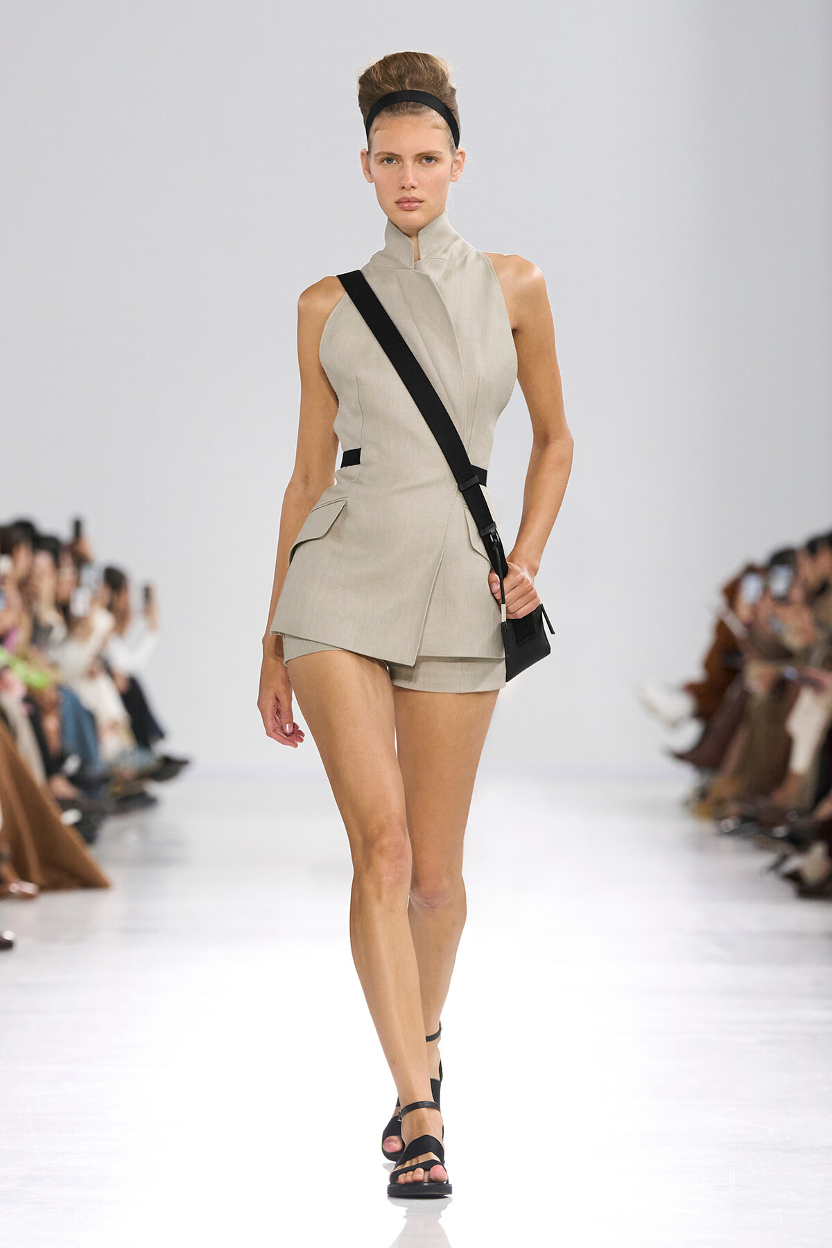 Model walks the runway in a beige sleeveless tailored romper with a high collar, paired with a black crossbody bag and headband.