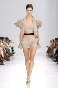 Model walks a runway in a beige outfit: strapless top, matching shorts, and a blazer with dramatic puff sleeves, cinched by a black belt, paired with black sandals.