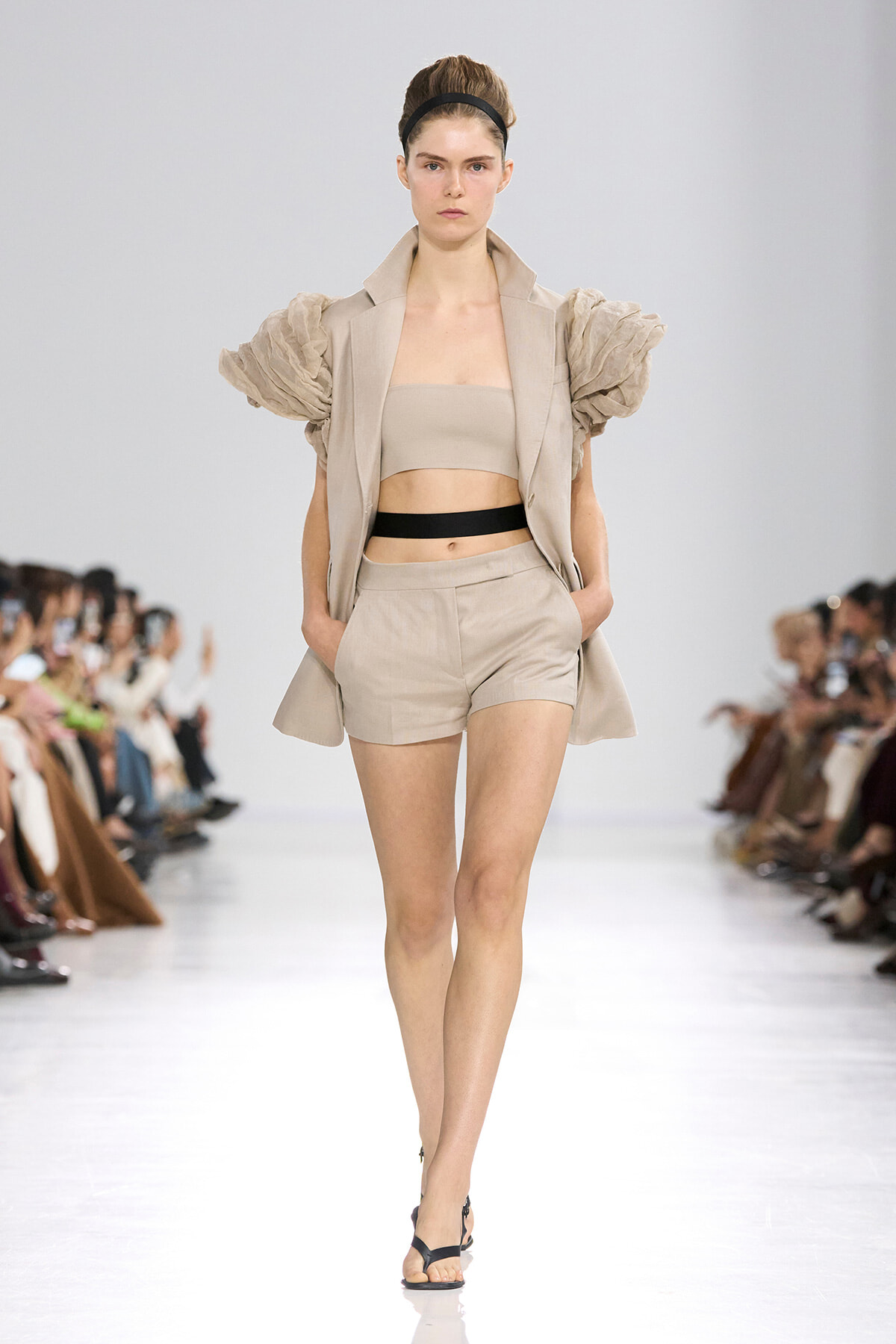 Model walks a runway in a beige outfit: strapless top, matching shorts, and a blazer with dramatic puff sleeves, cinched by a black belt, paired with black sandals.