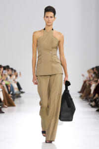 Model walks a runway in a taupe halter-neck double-breasted suit with wide-leg pants, carrying a black handbag.