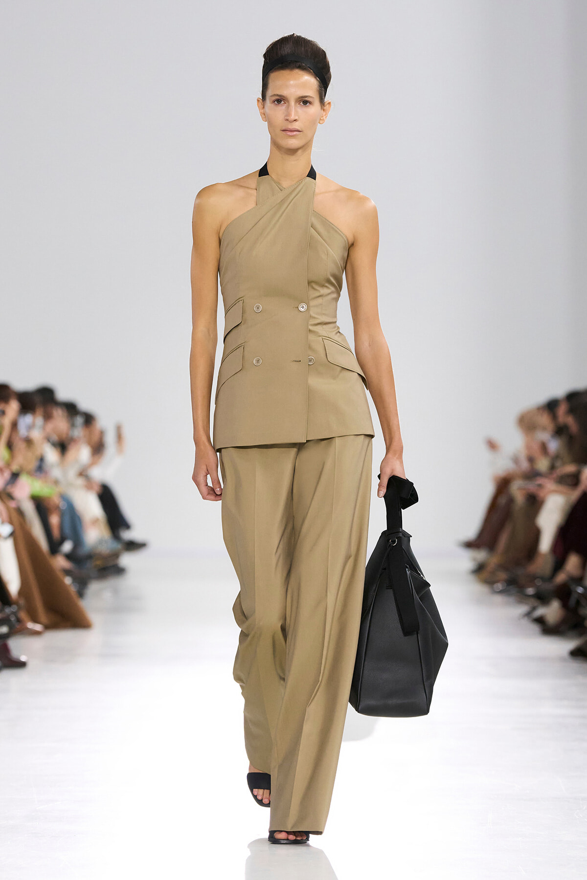Model walks a runway in a taupe halter-neck double-breasted suit with wide-leg pants, carrying a black handbag.