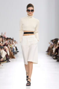 Model walking a runway in a cream cropped turtleneck sweater, a black waistband, and a white midi skirt with black sandals.