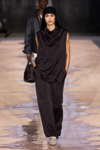 Model walking a fashion runway in a dark brown, draped sleeveless jumpsuit with a black headscarf and light-colored shoes, hands in pockets.
