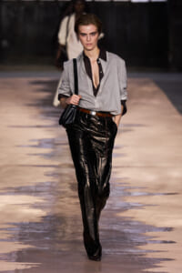 Model walking a runway in a light grey button-down shirt, black patent leather pants, brown belt, and a black shoulder bag.