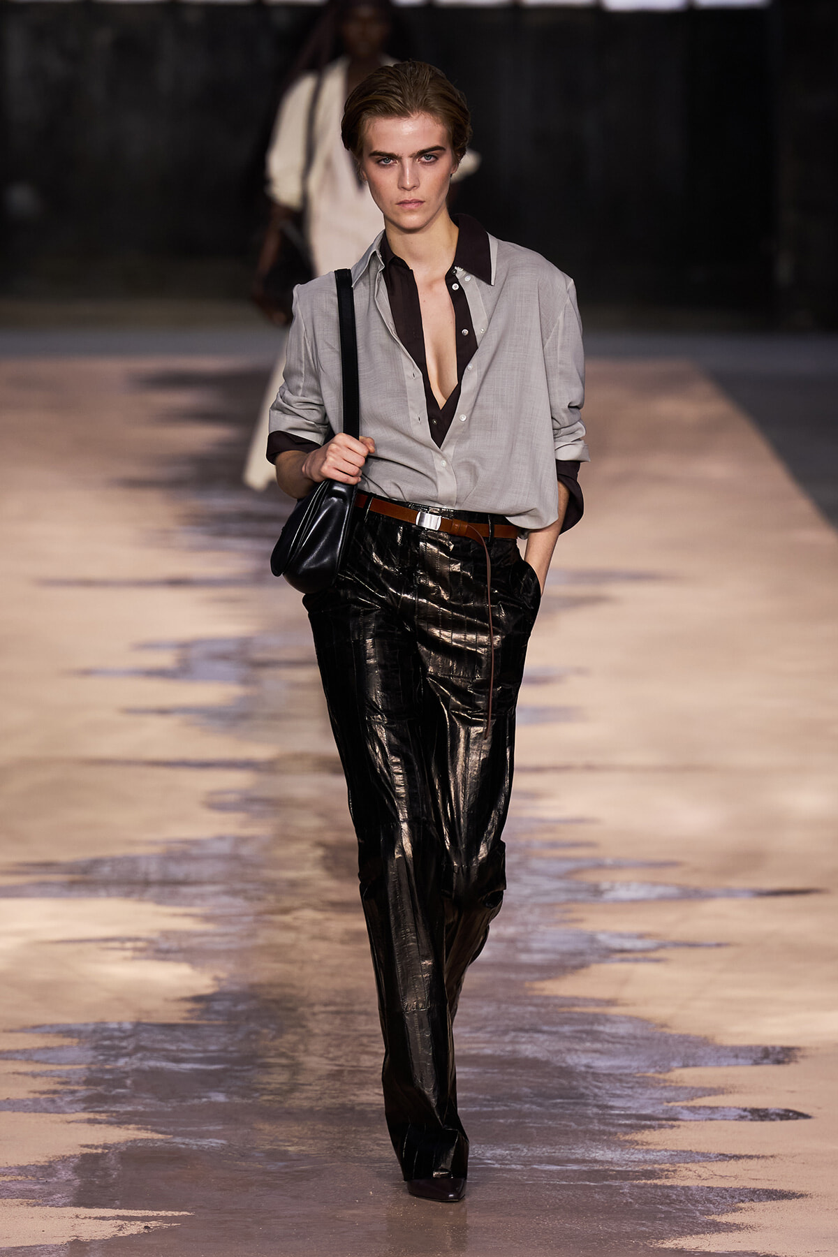 Model walking a runway in a light grey button-down shirt, black patent leather pants, brown belt, and a black shoulder bag.