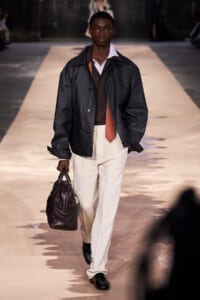 Male model on a runway wearing a dark jacket over a brown shirt, light beige trousers, an orange tie, and carrying a dark leather duffel bag.