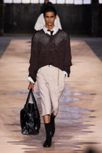 Male model walks a fashion runway in a dark knit sweater over a white shirt, beige cropped trousers, carrying a black leather tote.