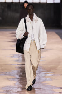Male model walking the runway in a white oversized jacket, striped shirt, and beige trousers, carrying a black shoulder bag.