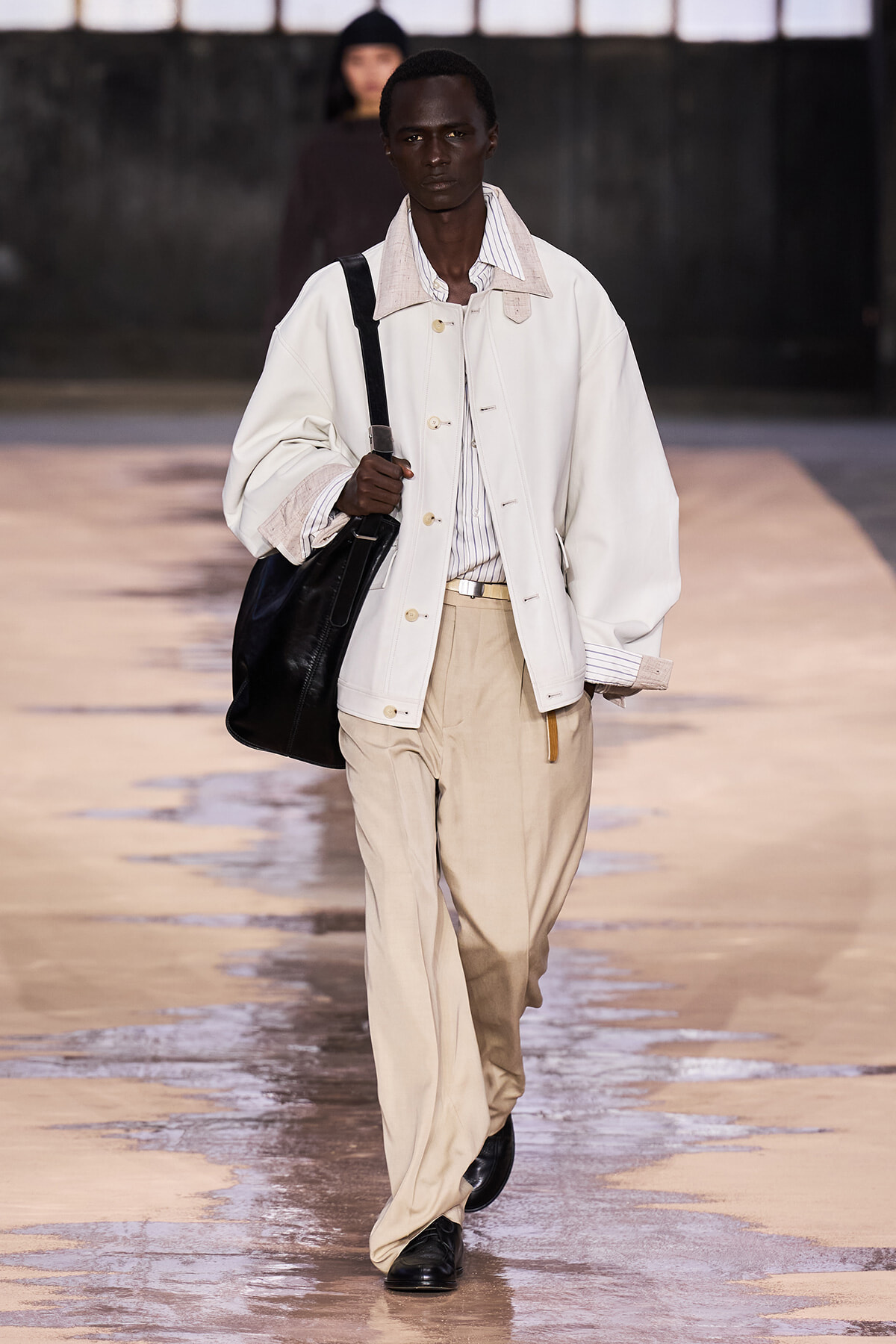 Male model walking the runway in a white oversized jacket, striped shirt, and beige trousers, carrying a black shoulder bag.