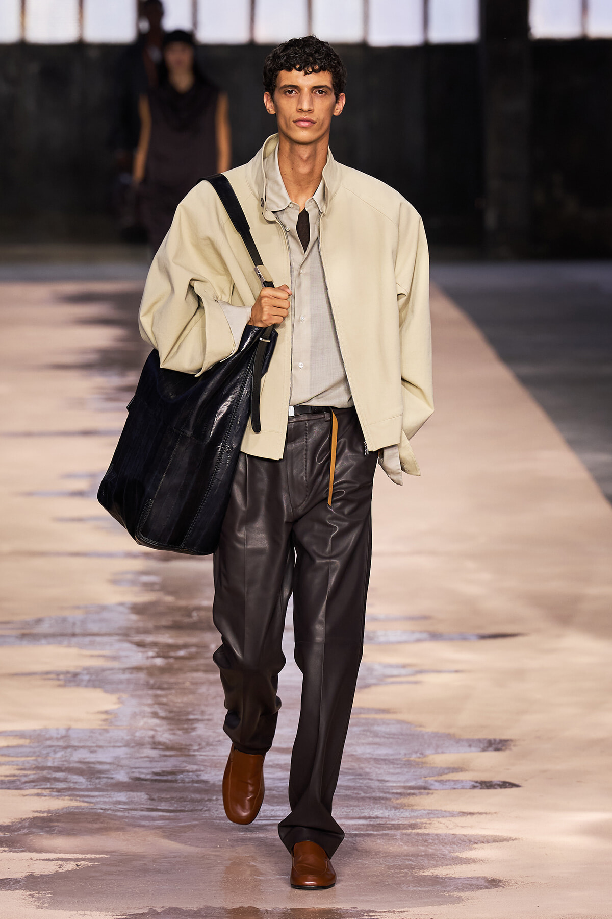 Male model walking a runway with a large black bag over the shoulder, wearing a beige jacket, gray shirt, dark leather pants, and brown shoes.
