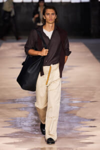 Male model walking a runway with a large black shoulder bag, wearing a dark shirt and beige cargo pants.
