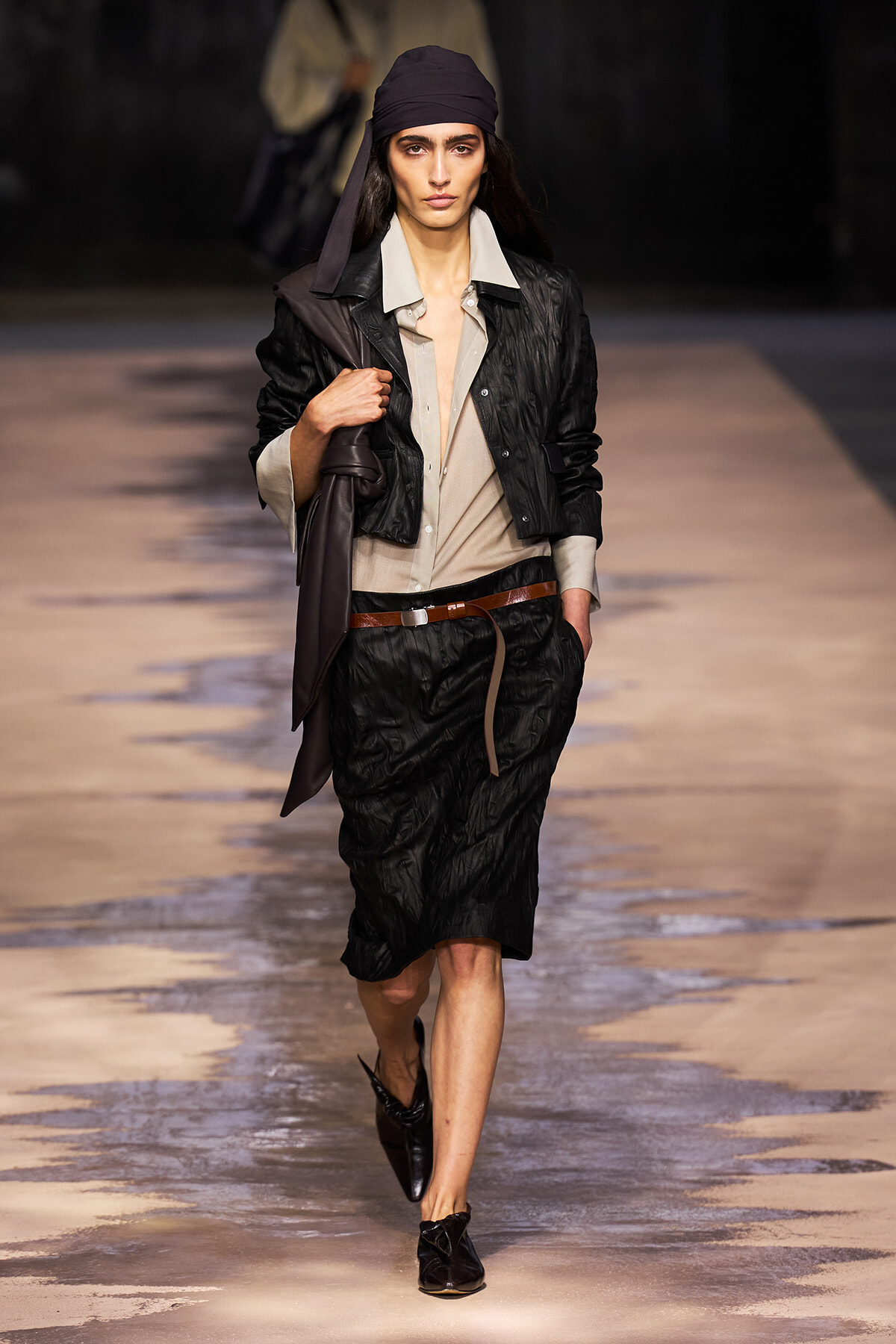 Model walking the runway in a black quilted suit with a beige button-up shirt, brown belt, and black headscarf, carrying a coat over the shoulder.