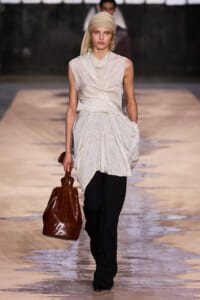 Model walking a runway in a sleeveless off-white draped tunic with black pants and a tan headscarf, carrying a brown leather handbag.