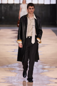 Male model walking a runway in a beige shirt, light tie, long black coat with brown cuffs, and black dress pants.