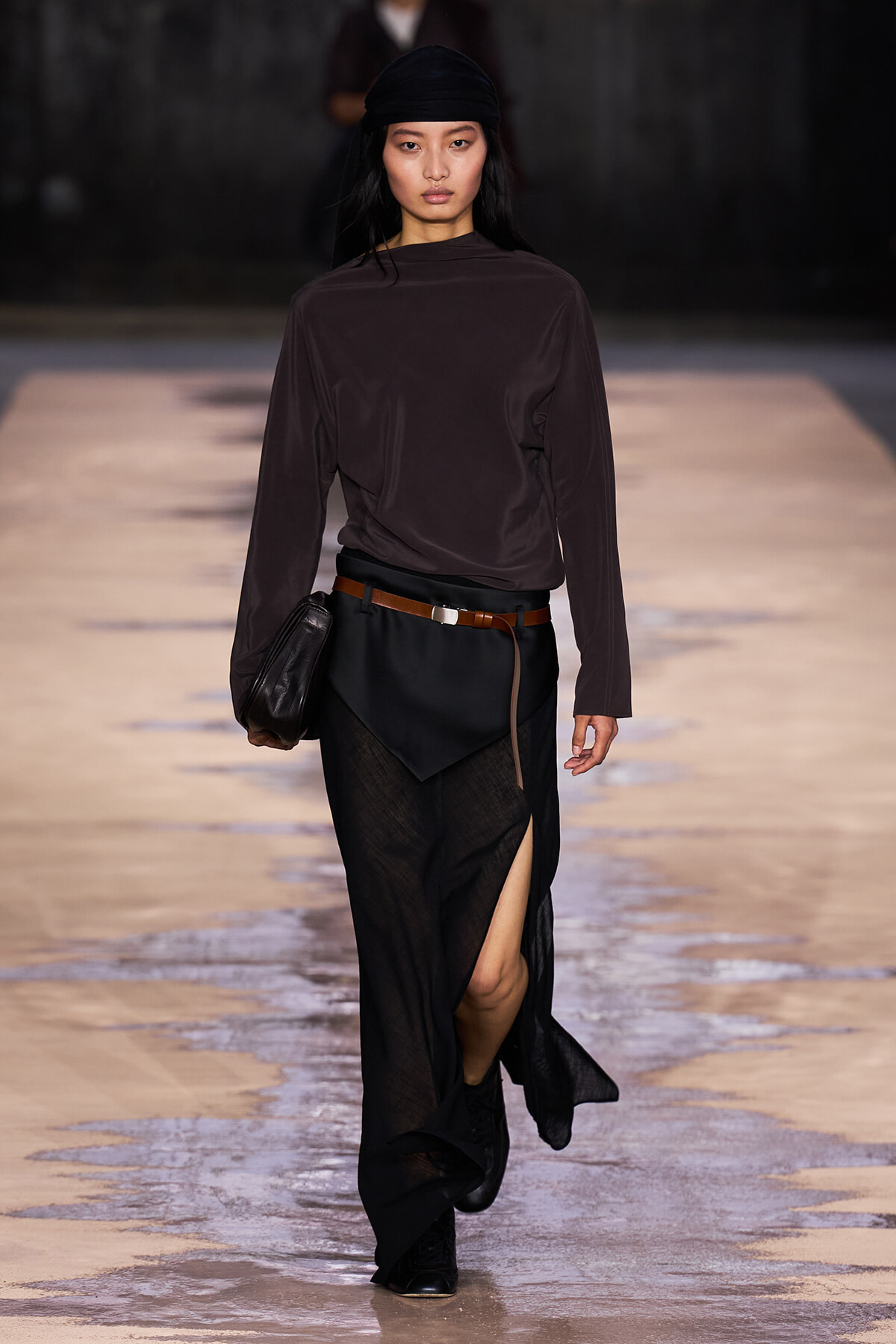 Model walking a runway in a dark brown long-sleeve top, black wrap skirt with a brown belt, and black boots, carrying a small black bag.