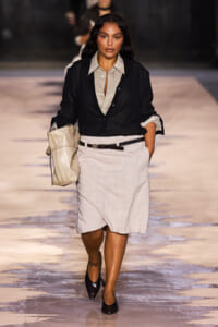 Model walking runway in a beige shirt with black cardigan and light beige knee-length wrap skirt, carrying a large beige handbag, wearing black heels.