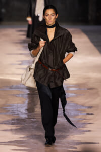 Model walking a runway in a loose dark brown jacket cinched with a brown belt, black pants, and a beige shoulder bag.