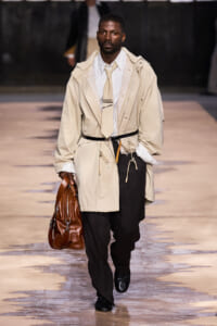 Male model walking runway in beige trench coat, white shirt, tan tie, black trousers, carrying brown leather bag.