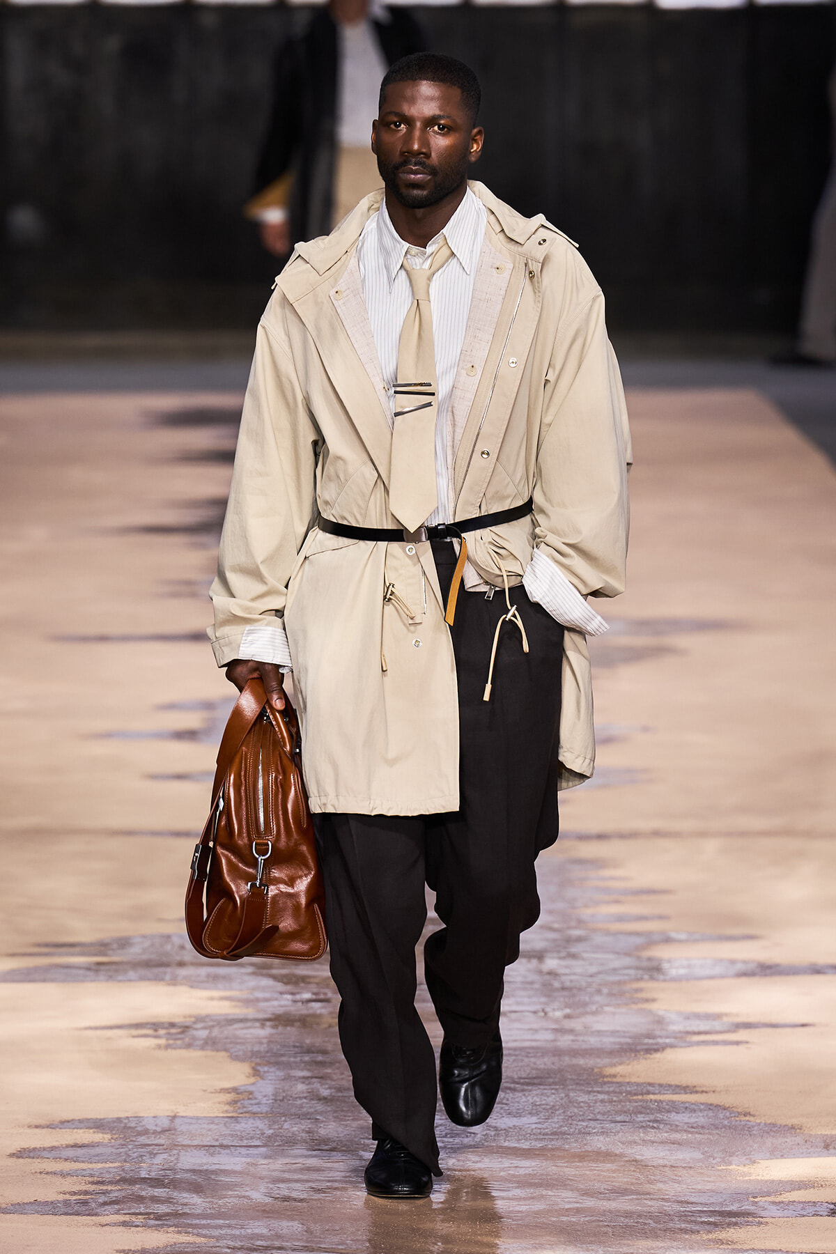 Male model walking runway in beige trench coat, white shirt, tan tie, black trousers, carrying brown leather bag.