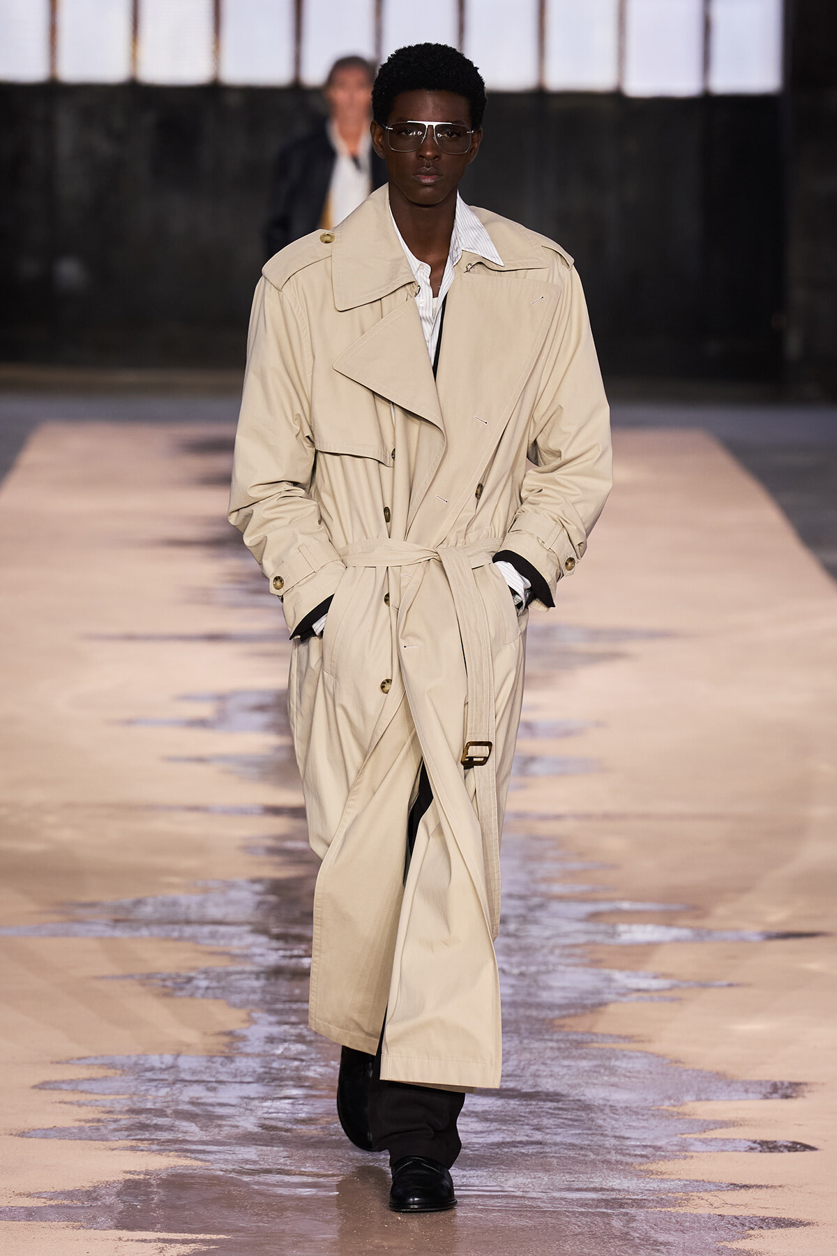 Model walks the runway in a long beige trench coat with a belt, black pants, and dark sunglasses on a glossy catwalk.