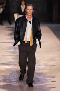 Male model on a runway wearing a black leather jacket over a white striped shirt with a tan scarf and dark brown pants.