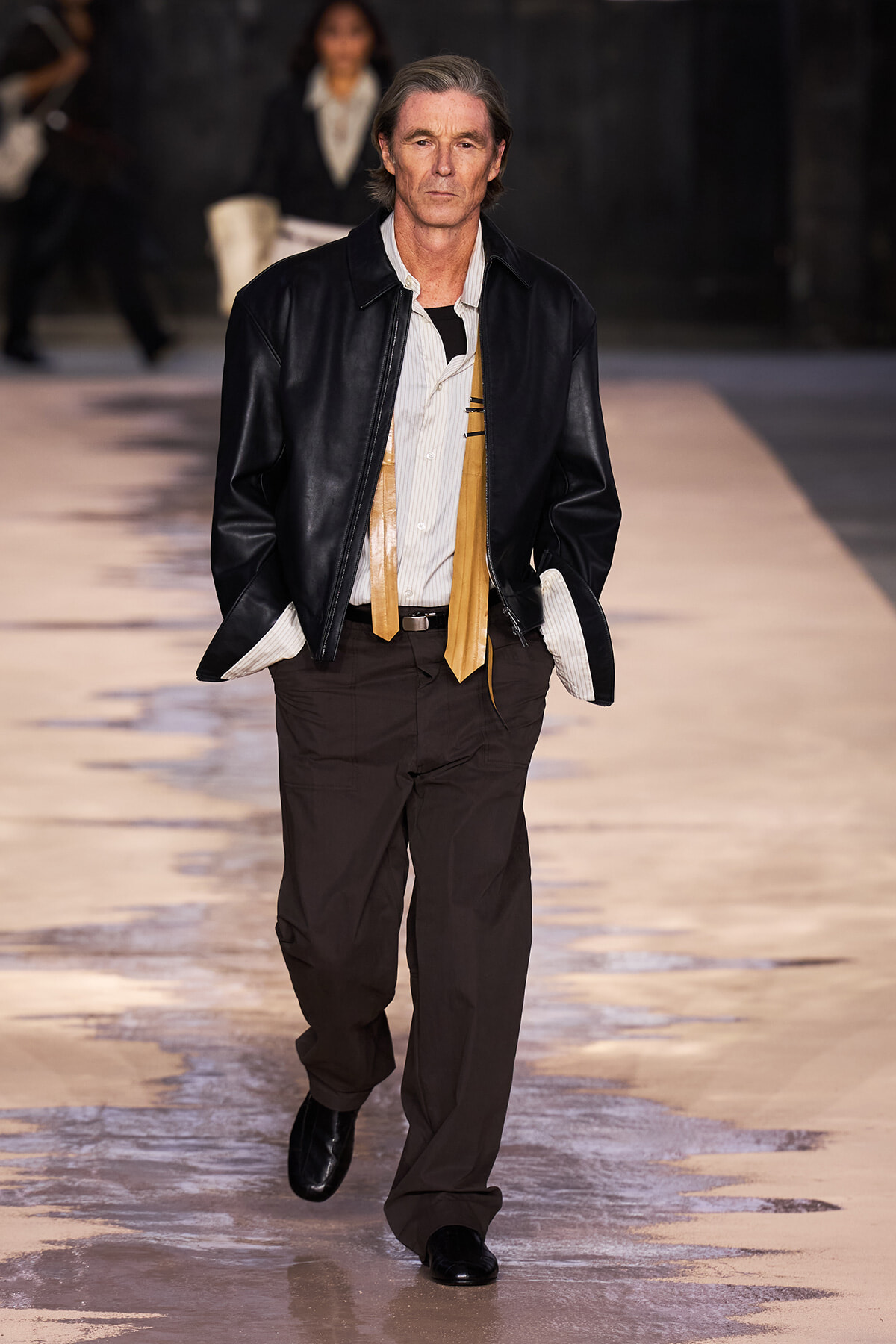 Male model on a runway wearing a black leather jacket over a white striped shirt with a tan scarf and dark brown pants.