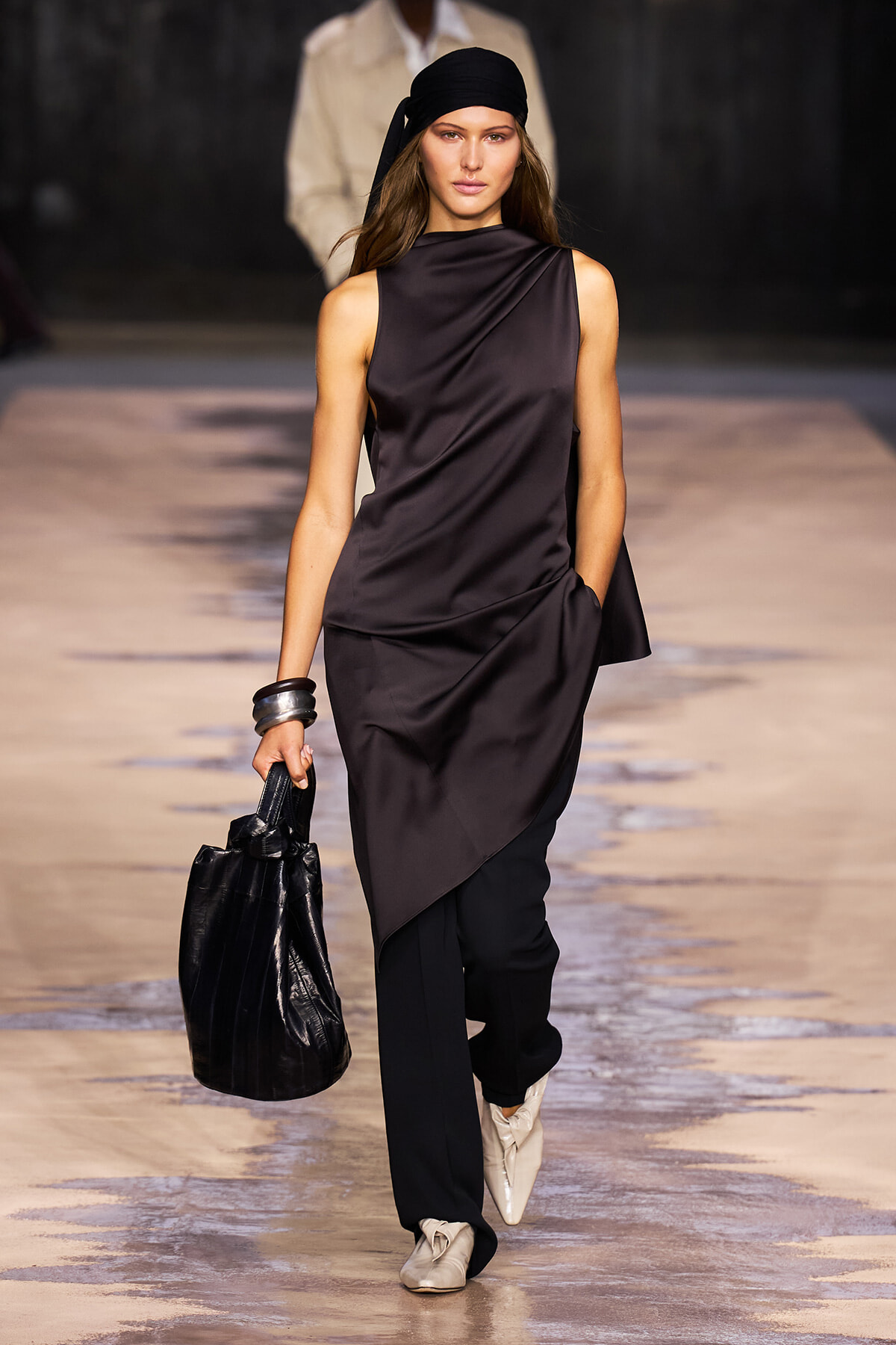 Model walks a fashion runway in a monochrome black satin draped outfit with a black headscarf, carrying a black pleated handbag and wearing light-colored ankle boots.