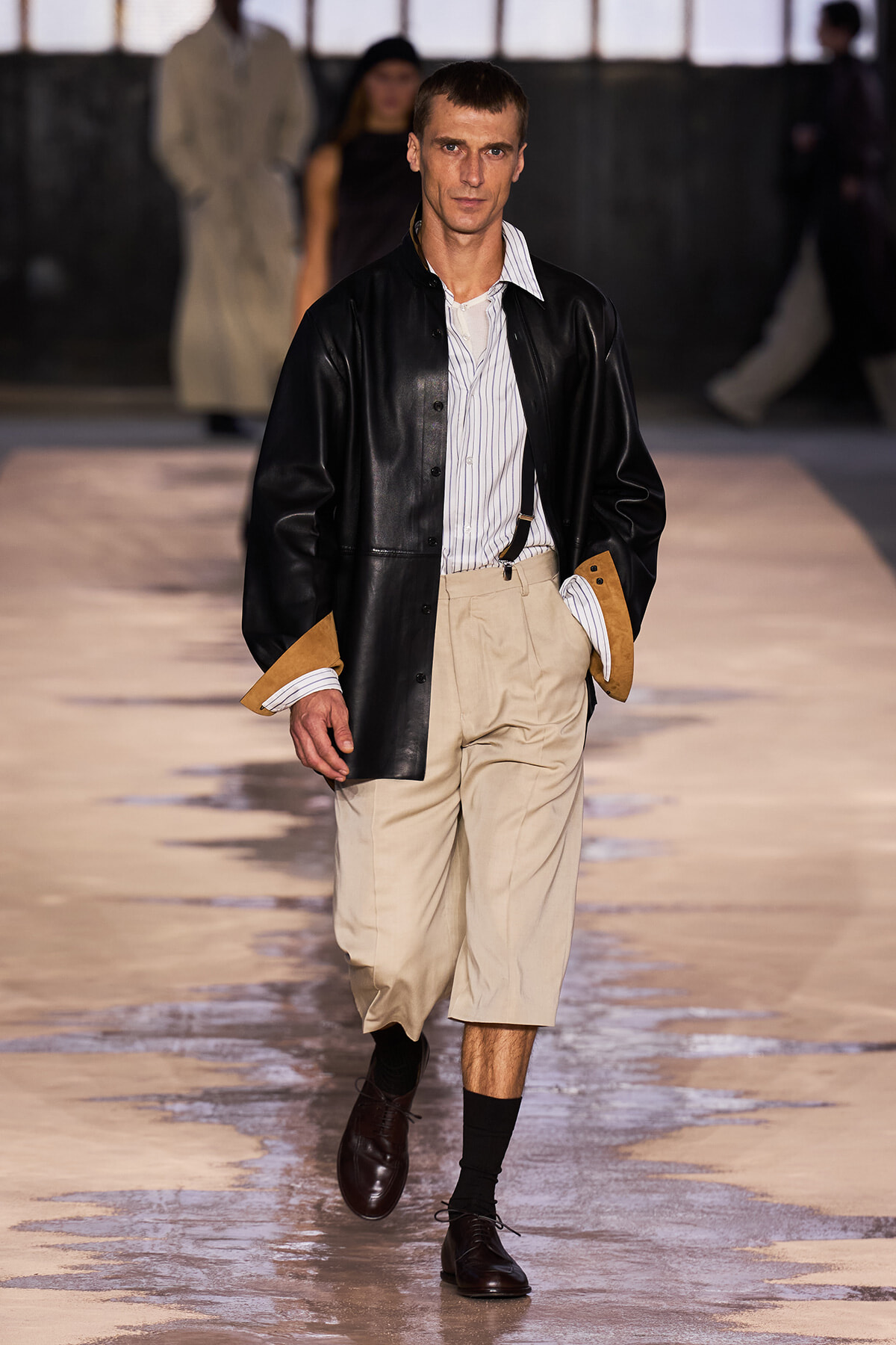Male model walking runway in black leather jacket, striped shirt, beige wide-leg pants, suspenders, and dark brown shoes.