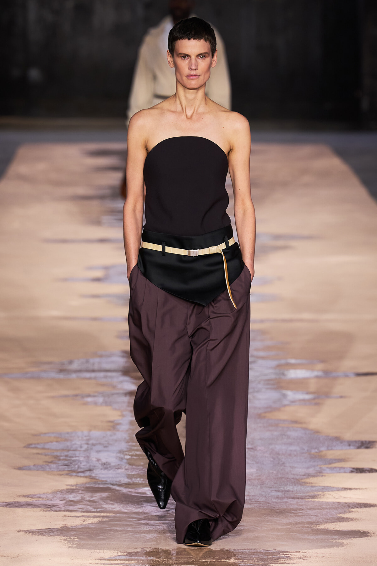 Model walks a fashion runway in a strapless black top with oversized maroon trousers and a beige belt.