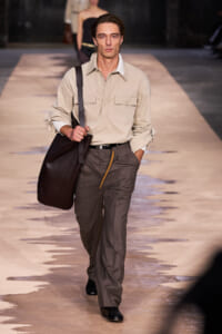 Male model walking a runway, wearing a beige utility shirt, gray trousers, and carrying a large dark bag over his shoulder.