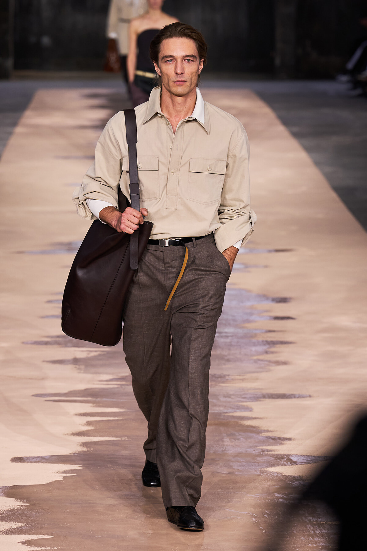 Male model walking a runway, wearing a beige utility shirt, gray trousers, and carrying a large dark bag over his shoulder.