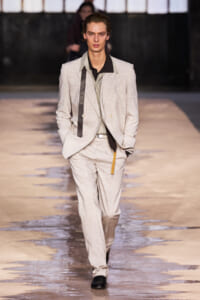 Male model walking a runway in a light beige suit with a black shirt, hands in pockets and a confident stride.