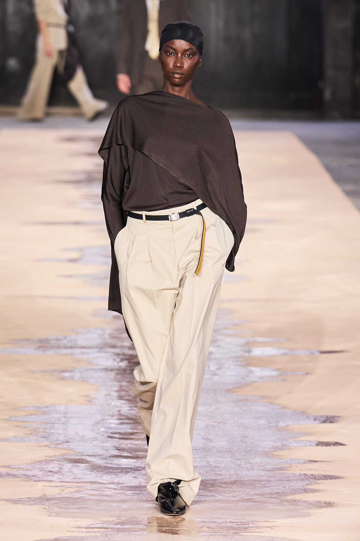 Model on a glossy runway wearing a dark brown draped top, cream wide-leg pants, black belt with a yellow hanging strap.
