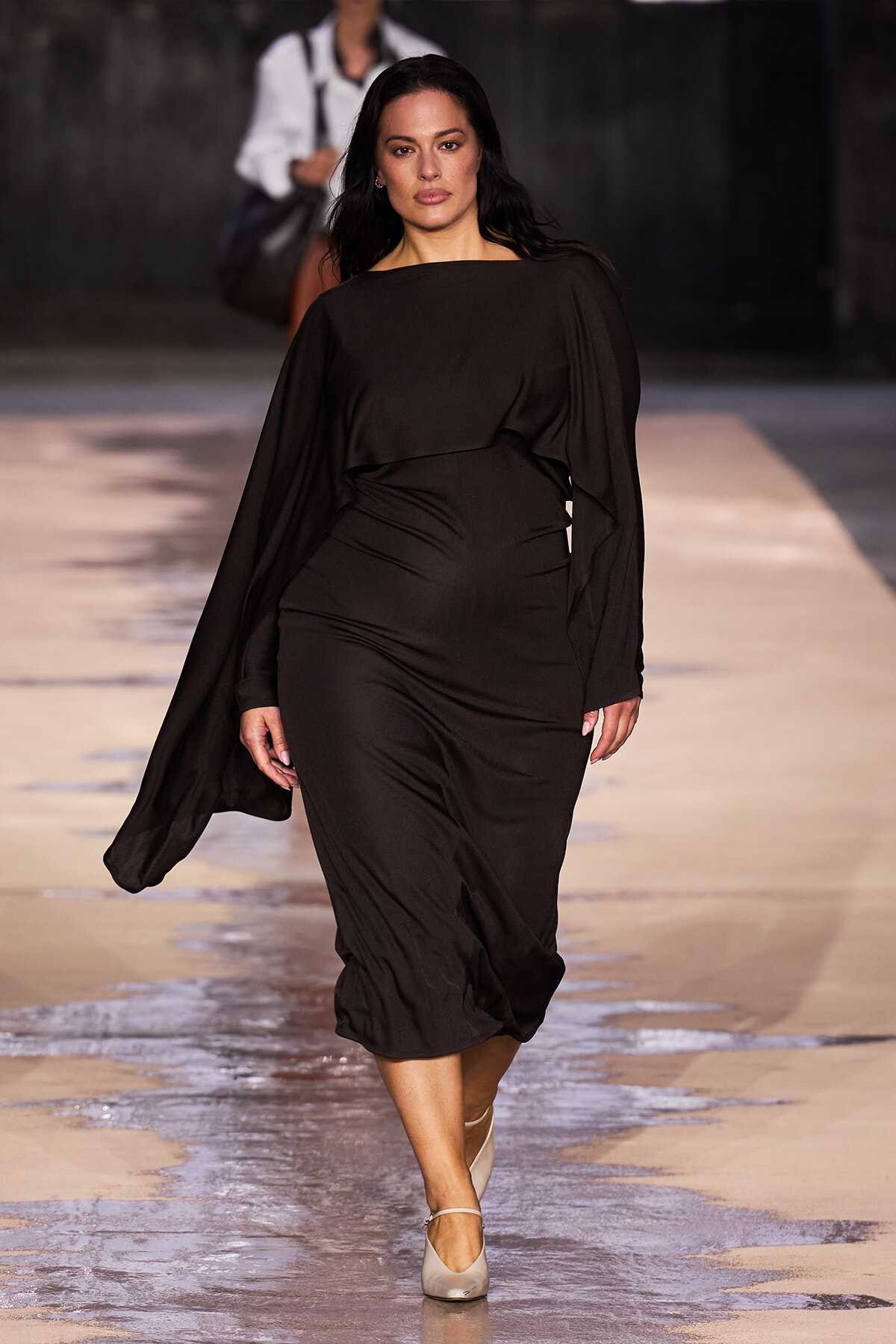 Plus-size model walks a fashion runway in a black draped dress with cape-like sleeves.