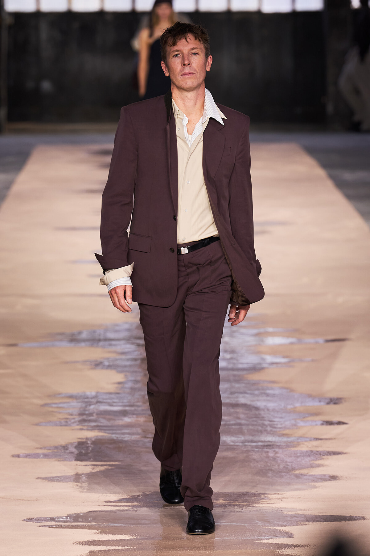 Male model walking down a fashion runway in a brown suit with a beige shirt, black belt and black shoes.