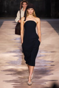 Female model walks a runway in a strapless black midi dress and brown headscarf, beige heels.