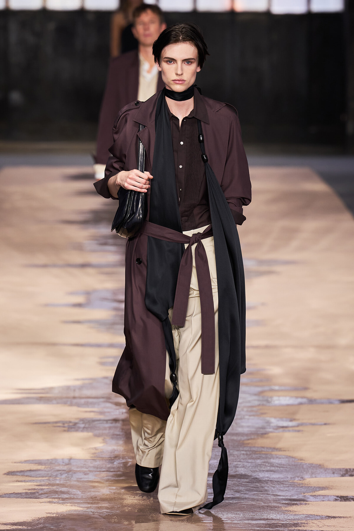 Model walking a fashion runway in a burgundy trench over a brown shirt with beige wide-leg pants, black scarf, and belt, carrying a black clutch.