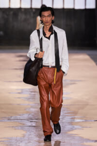 Male fashion model walking the runway in a white pinstriped shirt, brown leather pants, suspenders, and a black bag over the shoulder